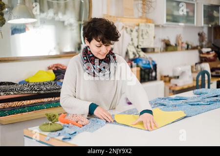 Konzentrierte lächelnde Designerin in lässiger Kleidung und Schal, die Muster auf Stoff vorbereitet, während sie am Tisch im Atelier arbeitet Stockfoto