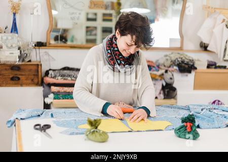 Konzentrierte lächelnde Designerin in lässiger Kleidung und Schal, die Muster auf Stoff vorbereitet, während sie am Tisch im Atelier arbeitet Stockfoto