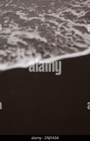Verschwommener Hintergrund von schaumigen Wellen mit Wellen, die sich am Sommertag am dunklen Sandstrand Rollen Stockfoto