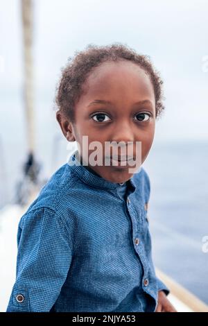 Positiver afroamerikanischer Junge in lässigem Hemd, der lächelt und in die Kamera schaut, während er auf einer Yacht gegen das Meer sitzt Stockfoto