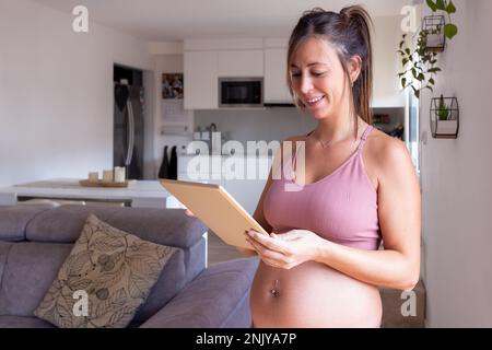Positive junge schwangere Frau in rosa Oberseite lächelt und surft im Internet auf einem Tablet, während sie zu Hause neben dem Sofa gegen die Küche steht Stockfoto