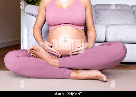 Verkürzte, nicht wiedererkennbare schwangere Frau in pinkfarbener Aktivkleidung und berührt den Bauch, während sie auf einer Yoga-Matte saß, in Lotus-Pose gegen das Sofa Stockfoto