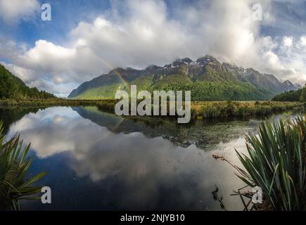 Malerischer Blick auf die Mirror Lakes, die den wolkigen blauen Himmel und Regenbogen in der Nähe der grünen Berge Neuseelands reflektieren Stockfoto