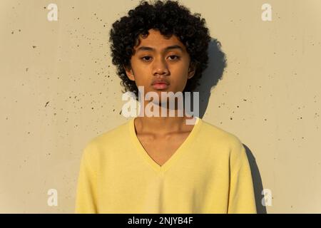 Selbstbewusster junger asiatischer Mann mit Afro-Frisur im gelben Sweatshirt, der vor der weißen Wand mit Schatten im Sonnenlicht auf die Kamera schaut Stockfoto