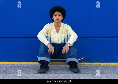 Ein ganzer Körper aus einem jungen, ethnischen Skater in lässiger Kleidung mit Afro-Frisur, sitzt auf dem Skateboard und sieht in der Ruhe in die Kamera Stockfoto