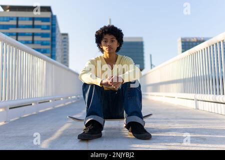 Der ganze Körper eines jungen asiatischen Skateboarders im lässigen Outfit sieht in die Kamera, während er auf einer Brücke auf dem Skateboard sitzt Stockfoto