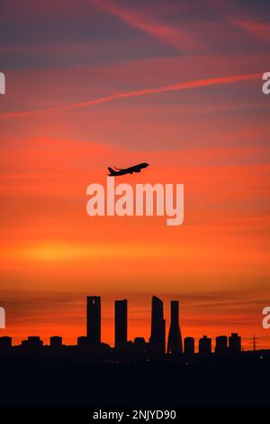 Silhouette des Flugzeugs, das über die Stadtlandschaft mit vier Türmen vor dem farbenfrohen Sonnenuntergang in Madrid fliegt Stockfoto