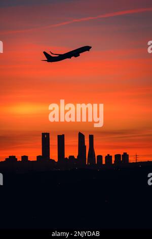 Silhouette des Flugzeugs, das über die Stadtlandschaft mit vier Türmen vor dem farbenfrohen Sonnenuntergang in Madrid fliegt Stockfoto