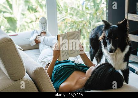 Süßer, neugieriger Border Collie Hund neben einer jungen Besitzerin, die auf einem bequemen Sofa liegt und am Wochenende zu Hause ein Interessantes Buch liest Stockfoto