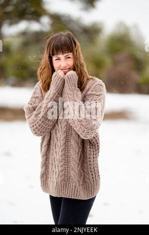 Positive junge Frau in warmem Strickpullover, die lächelt und in die Kamera schaut, während sie im Winterpark auf einem schneebedeckten Pfad steht Stockfoto