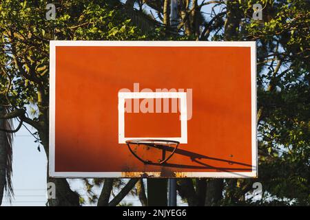 Der Basketballring hängt auf einer rostigen orangefarbenen und weiß bemalten Rückwand an hohen grünen Bäumen im Park auf der Straße und am blauen Himmel in Costa Rica Stockfoto