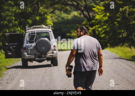 Rückansicht eines anonymen männlichen Reisenden in legerer Kleidung, der in Richtung eines SUV-Autos geht, das auf der Straße inmitten von Bäumen in Costa Rica geparkt ist Stockfoto