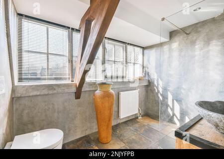 Innendesign des Badezimmers mit Glasdusche und weißer Toilette neben Waschbecken auf Holzwaschtisch und großer Vase neben Heizkörper an grauen Wänden Stockfoto