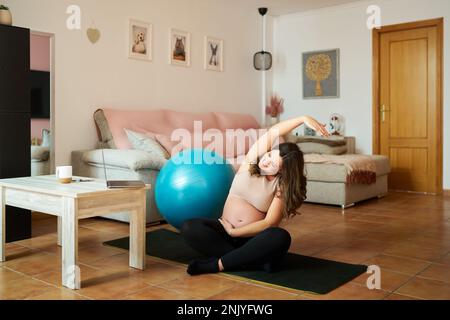 Der ganze Körper einer lächelnden jungen Frau in schwarzen Leggings, die Yoga für schwangere Frauen auf der Matte neben dem blauen Ball im Wohnzimmer macht Stockfoto