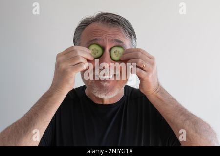 Grauer, lächelnder Mann, der die Augen im Studio mit Gurkenscheiben auf weißem Hintergrund bedeckt Stockfoto