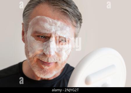 Positiv bärtiger älterer Mann, der eine Tonmaske auf dem Gesicht aufträgt, während er lächelt und während der Hautpflege gegen die weiße Wand in den Spiegel schaut Stockfoto