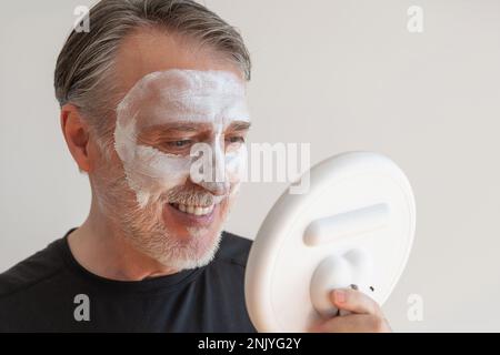 Positiv bärtiger älterer Mann, der eine Tonmaske auf dem Gesicht aufträgt, während er lächelt und während der Hautpflege gegen die weiße Wand in den Spiegel schaut Stockfoto