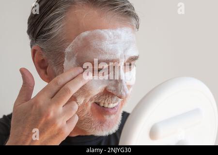 Positiv bärtiger älterer Mann, der eine Tonmaske auf dem Gesicht aufträgt, während er lächelt und während der Hautpflege gegen die weiße Wand in den Spiegel schaut Stockfoto