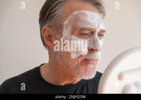 Positiv bärtiger älterer Mann, der eine Tonmaske auf dem Gesicht aufträgt, während er lächelt und während der Hautpflege gegen die weiße Wand in den Spiegel schaut Stockfoto