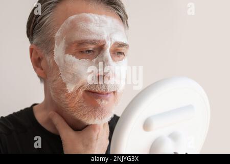 Positiv bärtiger älterer Mann, der eine Tonmaske auf dem Gesicht aufträgt, während er lächelt und während der Hautpflege gegen die weiße Wand in den Spiegel schaut Stockfoto