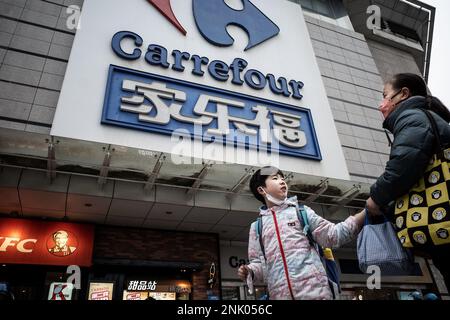 Ein Junge und eine Frau gehen an einem riesigen Schild von Carrefour in Wuhan vorbei. Carrefour trat im Jahr 1995 in den chinesischen Markt ein und expandierte sehr schnell, um einen großen Marktanteil zu sichern. Die Einnahmen nahmen jedoch mit zunehmender Konkurrenz ab. Im Jahr 2019 verkaufte der französische Einzelhändler rund 80 % seines „verlustbringenden“ Geschäftsbereichs in China für 4,8 Mrd. CNY an das in Jiangsu ansässige Unternehmen „Suning“, was zu diesem Zeitpunkt 699 Mio. USD entsprach. In diesem Monat haben Berichte über Engpässe in den zahlreichen lokalen Carrefour-Filialen die Aufmerksamkeit der Kunden erregt. Einige Leute begannen sich darüber zu beschweren Stockfoto