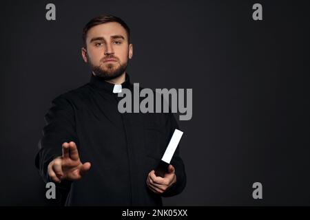 Priester mit Bibel macht Segen auf schwarzem Hintergrund. Platz für Text Stockfoto