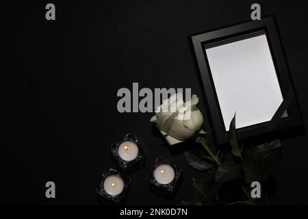 Bilderrahmen für die Beerdigung mit schwarzem Band, weißer Rose und Kerzen auf dunklem Hintergrund, flach liegend. Platz für Text Stockfoto