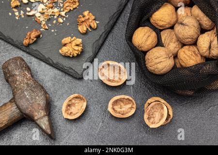 Walnüsse in einem wiederverwendbaren Beutel. Hammer, Mutternschalen auf dem Tisch und Nusskerne auf der Steinplatte. Schwarzer Hintergrund. Stockfoto