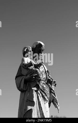 Eine getragene und verwitterte Natursteinstatue des Heiligen Joseph, die das Jesuskind in den Händen trägt, in Schwarz-Weiß-Schwarz. Stockfoto