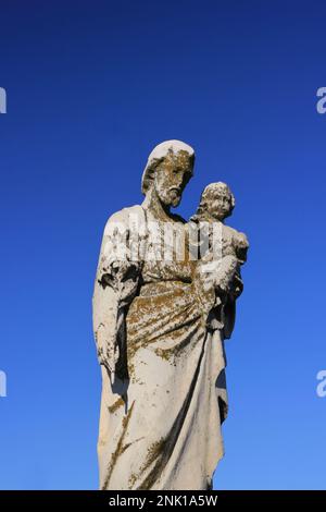 Eine getragene und verwitterte Natursteinstatue des Heiligen Joseph, die das Jesuskind in seinen Händen trägt. Stockfoto