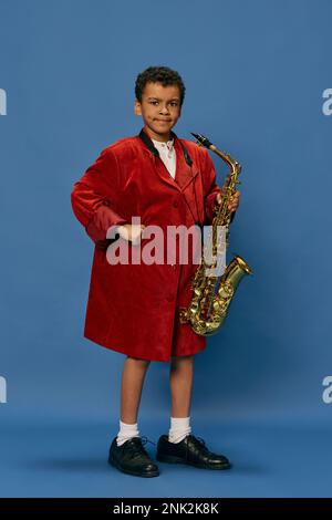 Portrait eines süßen, kleinen afrikanischen Jungen, der eine riesige Männerjacke trägt und Schuhe trägt, wie ein Jazz-Mann, der auf Saxofon spielt, vor blauem Hintergrund. Mode, Kunst, Musik Stockfoto