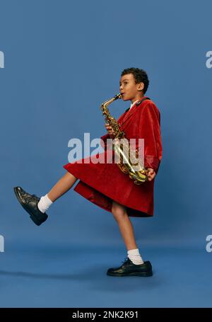 Portrait eines süßen, kleinen afrikanischen Jungen, der eine riesige Männerjacke trägt und Schuhe trägt, wie ein Jazz-Mann, der auf Saxofon spielt, vor blauem Hintergrund. Mode, Kunst, Musik Stockfoto