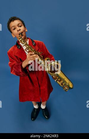Weitwinkelansicht eines süßen kleinen afrikanischen Jungen, der eine riesige Männerjacke trägt und Schuhe trägt, wie ein Jazzmann, der auf Saxophon spielt, vor blauem Hintergrund. Mode, Kunst Stockfoto