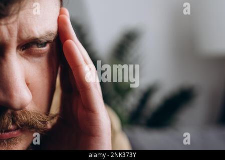 Gekürztes Porträt eines traurigen jungen Kaukasiers, der unter Migräne leidet, während er allein in der Wohnung ist. Unglückliche, traurige männliche Massagebügel, mentale Stres Stockfoto