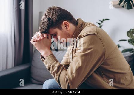 Nahaufnahme eines stressigen, traurigen jungen Mannes, der allein auf dem Sofa im Wohnzimmer sitzt und sein Gesicht mit Händen bedeckt. Depression männlicher 30s-Jähriger, verärgert und unh Stockfoto