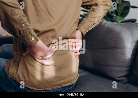 Seitenansicht eines ungesunden jungen Kaukasier, der zu Hause auf der Couch sitzt und unter Rückenschmerzen leidet, Rückenschmerzen, Gesundheitsprobleme, unterer Rücken, hat chronisch Stockfoto