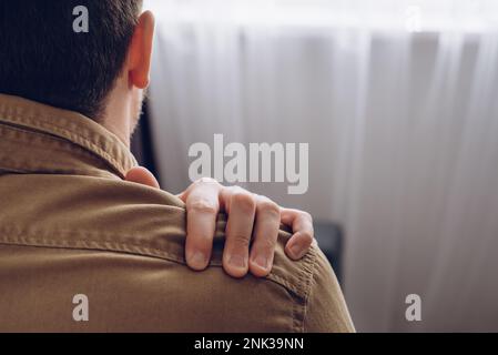Seitenansicht eines ungesunden jungen Mannes, der in der Nähe des Fensters sitzt und unter Schmerzen in der Schulter leidet und Selbstmassage durchführt. Nackenschmerzen. Unterarmschmerzen, Menschen mit Körper mu Stockfoto