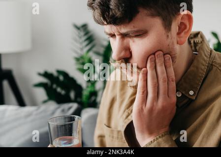 Trauriger weißer Mann 30s sitzt zu Hause auf der Couch und berührt die Wange, fühlt sich verletzt und leidet an empfindlichen Zahnschmerzen. Schmerz- und Hohlraumkonzept. Aufgebracht, jung Stockfoto