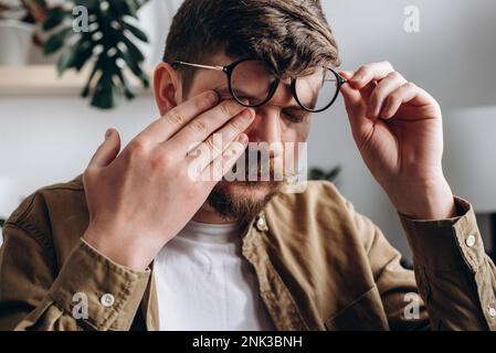 Nahaufnahme eines verärgerten jungen bärtigen Mannes mit Brille, der Nasenbrücke reibt, sitzen auf dem Sofa im Büro. Trauriger Weißer, männlich, 30s, berührt das Gesicht, will schlafen, f Stockfoto