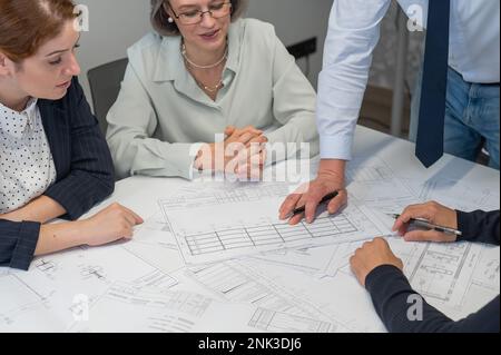 Ein weißer Mann steht und ändert eine Zeichnung, drei Kollegen sitzen an einem Tisch und hören ihm zu. Stockfoto