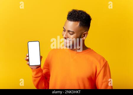 Attraktiver junger Mann, der ein Smartphone mit leerem Bildschirm zeigt und auf den Bildschirm schaut, der Kerl mag eine mobile App, fühlt sich zufrieden, isoliert Stockfoto