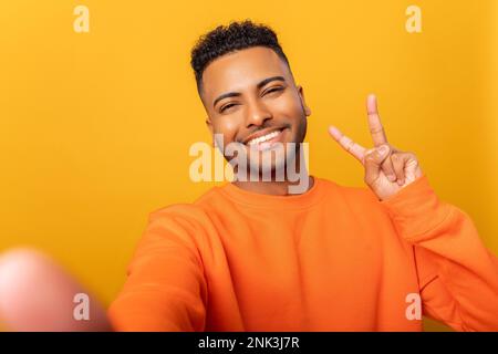 Glücklicher zufriedener Mann, der das V-Zeichen des Friedens mit den Fingern zeigt, mit einem Zahnlächeln in die Kamera schaut, Selfie macht. Innenstudio-Aufnahme isoliert auf gelbem Hintergrund Stockfoto