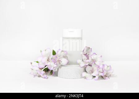 Durchsichtiges Mikellarwasser in einer weißen Plastikflasche auf einem Podiumsockel auf weißgrauem Hintergrund mit Blume, Pflanze. Make-up-Reiniger für Gesichtshaut. Stockfoto