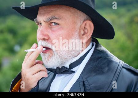 Nahaufnahme Außenporträt von stilvoller Mode, alter alter alter alter alter Senior in Ledermantel und modischer Mütze. Alter Mann, der raucht. Stockfoto