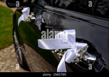 Dekorationen und Details auf dem Hochzeitsauto weißes Band und winzige Blumen am Türgriff eines schwarzen, modernen Autos kleine Feldtiefe Stockfoto