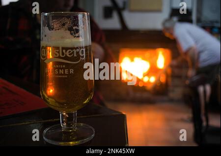 Ein Pint Carlsberg-Lager an der Bar im Pub mit einem brennenden Kaminfeuer im Hintergrund Stockfoto