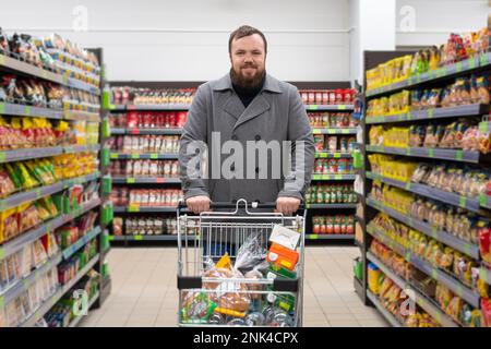 Ein lächelnder, zufriedener junger Mann mit einem Bart und einem grauen Mantel steht in einem Supermarkt mit einem mit Essen gefüllten Einkaufswagen Stockfoto