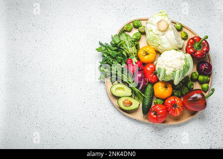 Rustikales Holztablett mit Auswahl an frischem Gemüse und Grün auf weißem Küchentisch mit Blick von oben. Vegetarische und diätetische Lebensmittel aus biologischen Zutaten Stockfoto