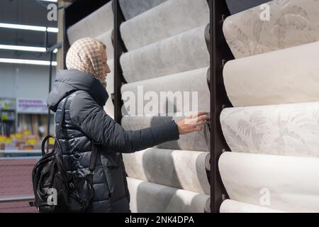 Eine junge Frau in einem Daunenmantel wählt eine helle Tapete in einem Baumarkt. Konzeption und Rekonstruktion des Hauses Stockfoto