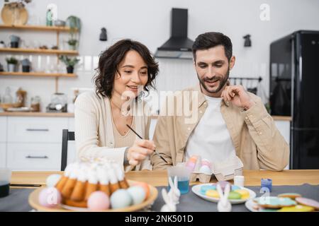 Ein positives Paar malte Eier in der Nähe von verschwommenen Ostereiern in der Küche Stockfoto